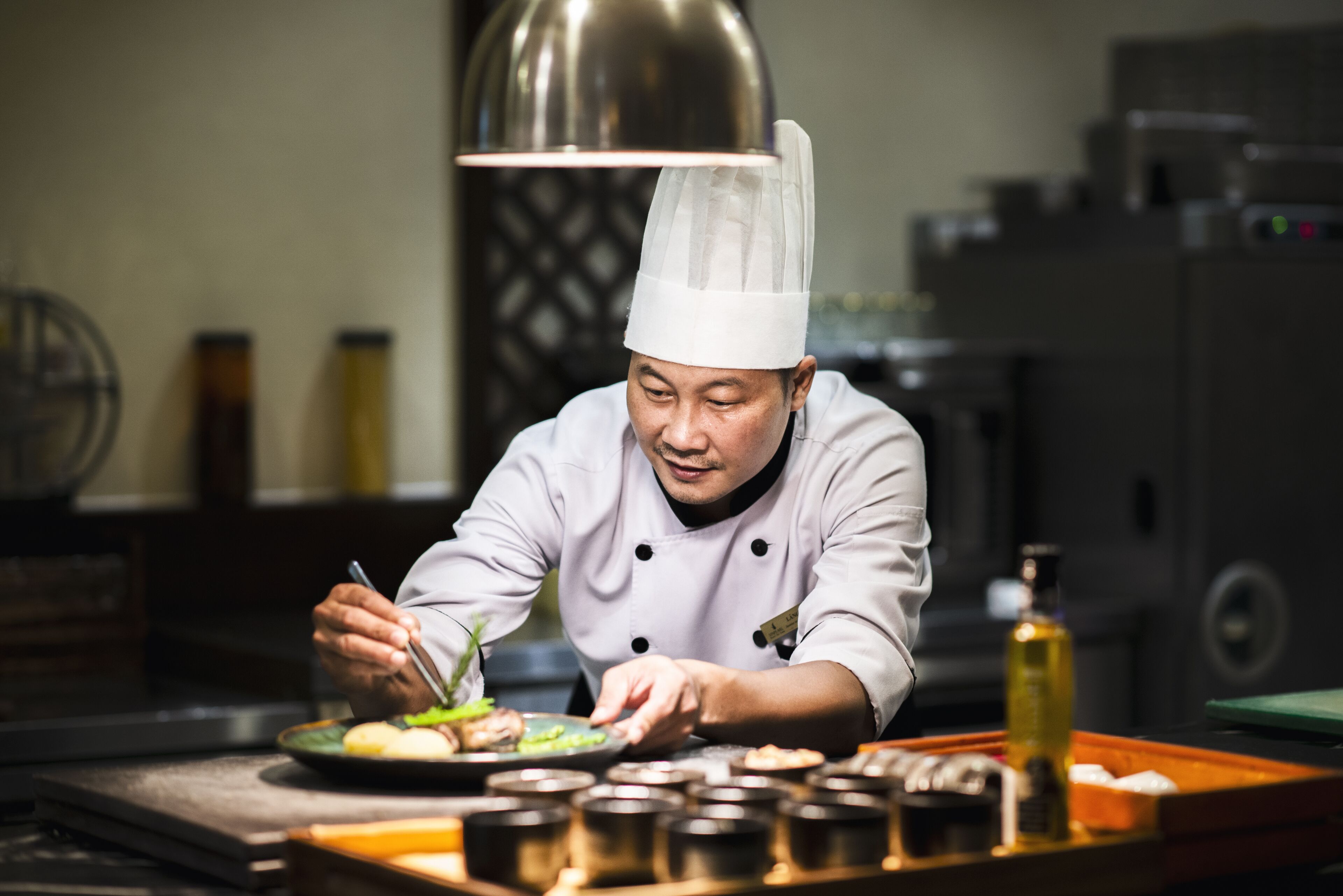 Un chef, vêtu d’un uniforme blanc et coiffé d’un haut-de-forme, prépare des plats dans une cuisine éclairée par un grand luminaire suspendu, où l’on peut voir divers ustensiles de cuisine et ingrédients disposés sur le plan de travail.