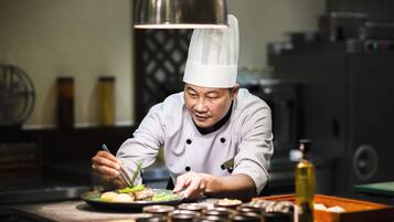 Un chef, vêtu d’un uniforme blanc et coiffé d’un haut-de-forme, prépare des plats dans une cuisine éclairée par un grand luminaire suspendu, où l’on peut voir divers ustensiles de cuisine et ingrédients disposés sur le plan de travail.