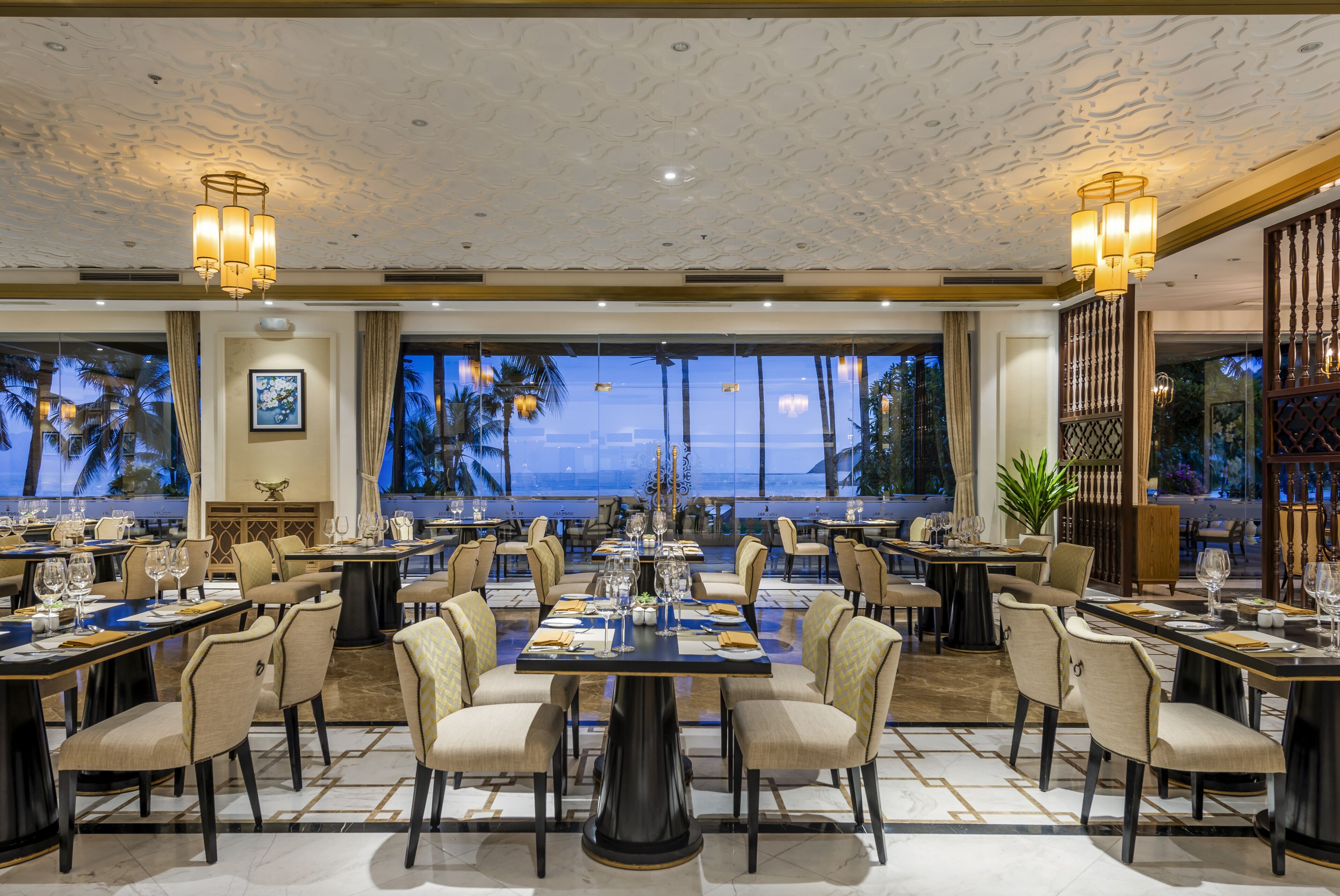 A spacious dining area with multiple tables and chairs, large windows with a view of palm trees, and a ceiling with a textured pattern.