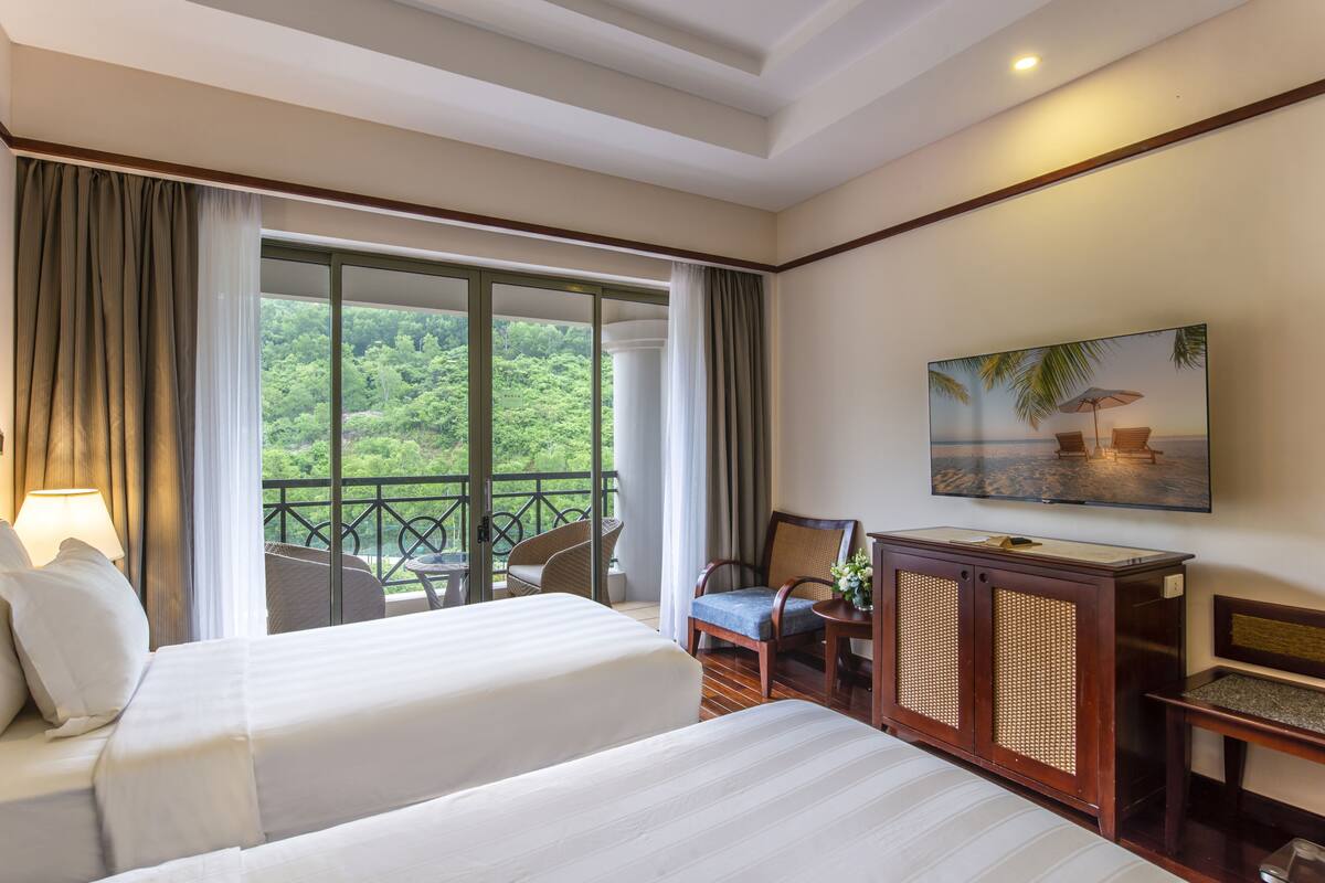 Een hotelkamer met twee bedden, een balkon met uitzicht op groen, een televisie en een houten kast.
