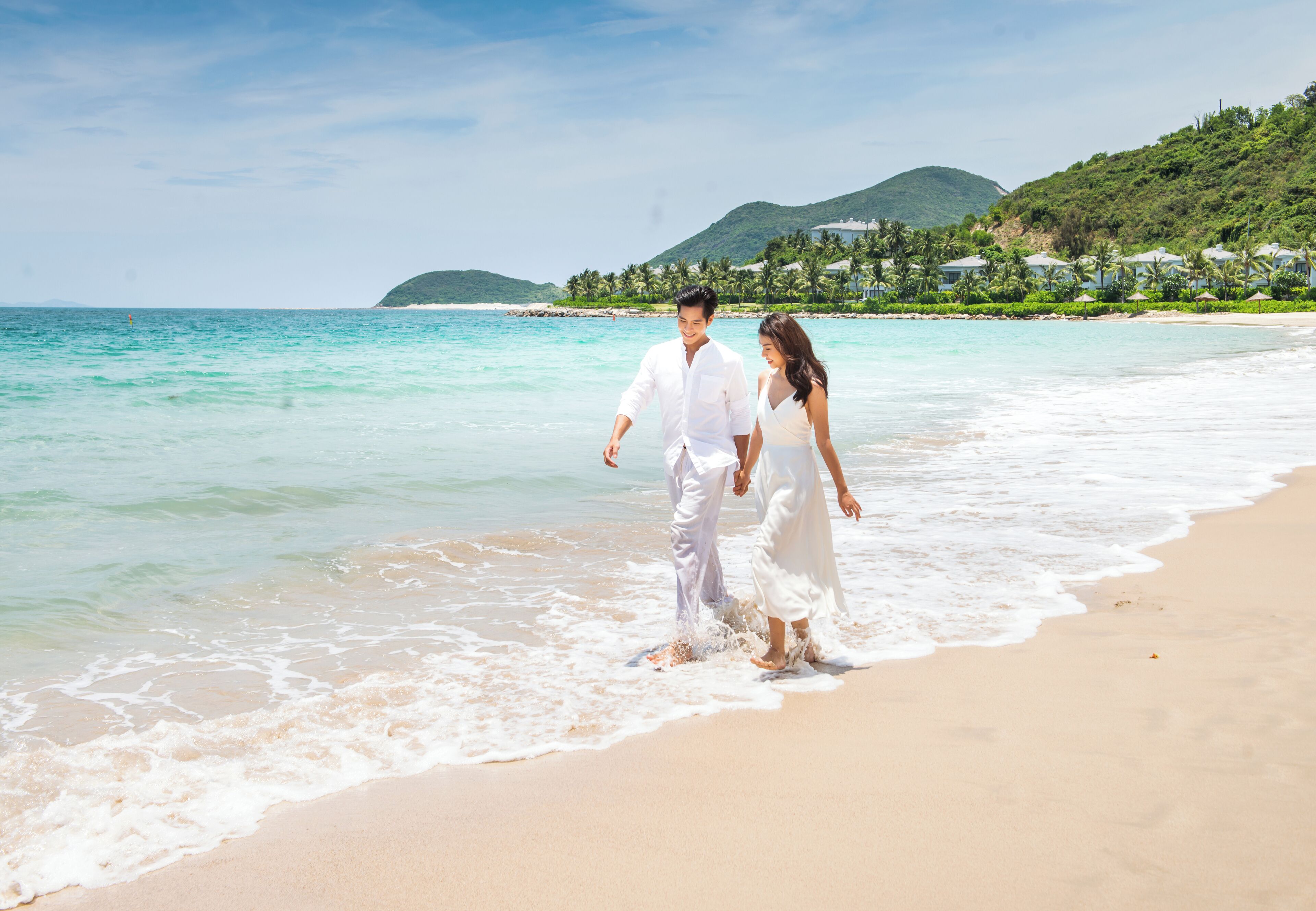 Um casal caminhando numa praia com águas turquesa cristalinas e montanhas ao fundo.