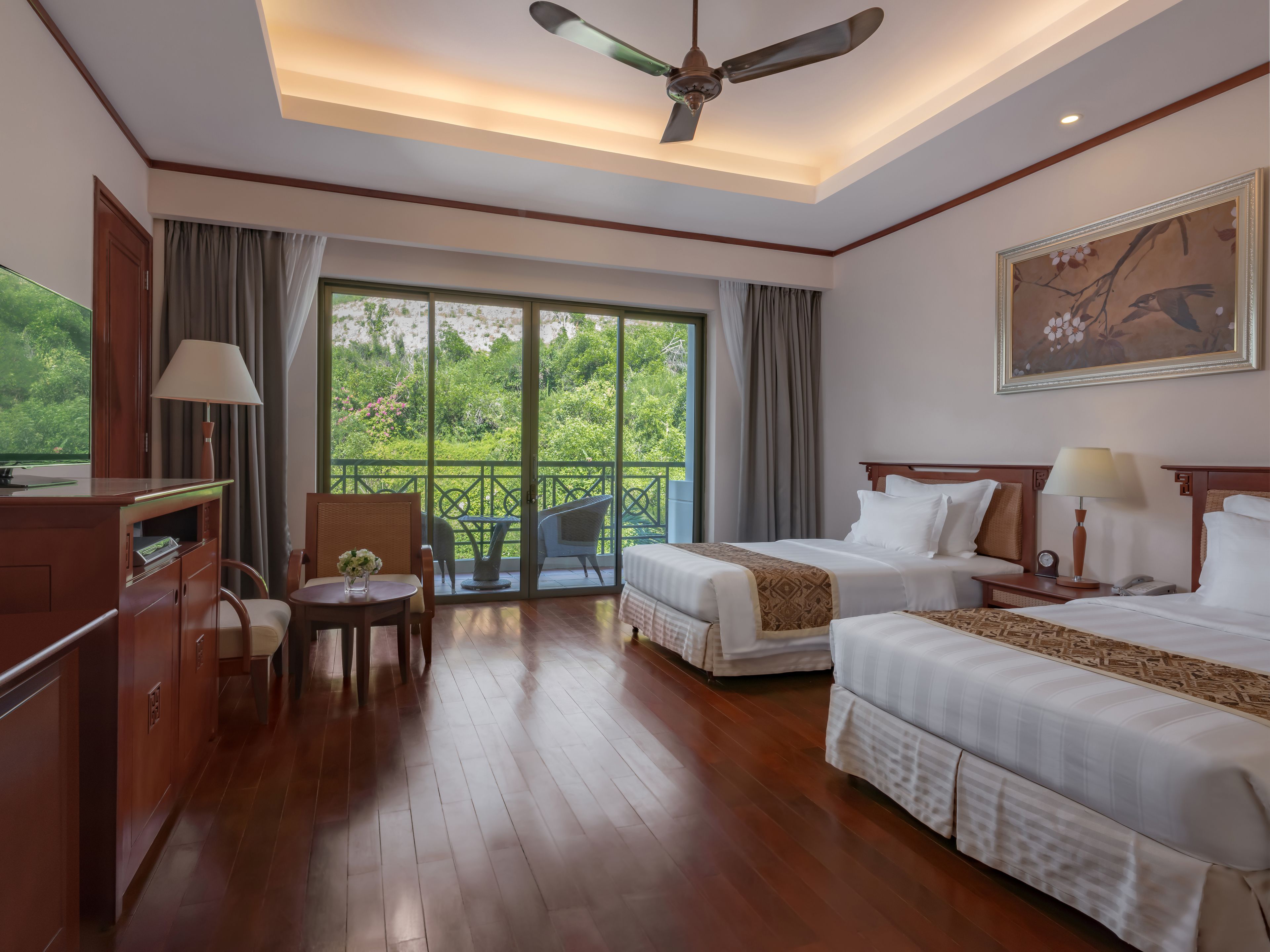 A hotel room with two beds, a desk, a chair, a table with a lamp, and a balcony with a view of greenery.