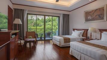 A hotel room with two beds, a desk, a chair, a table with a lamp, and a balcony with a view of greenery.