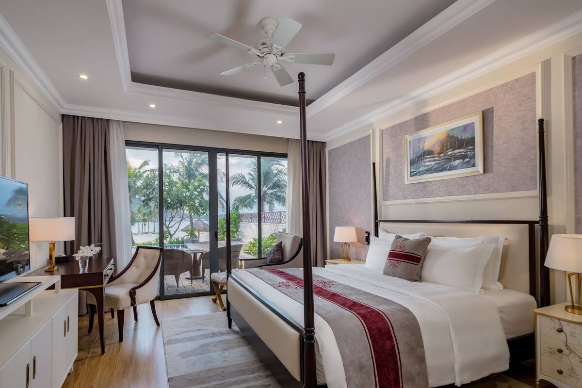 Hotelkamer met een bed, een bureau, een stoel, een televisie en uitzicht op palmbomen en een zwembad.