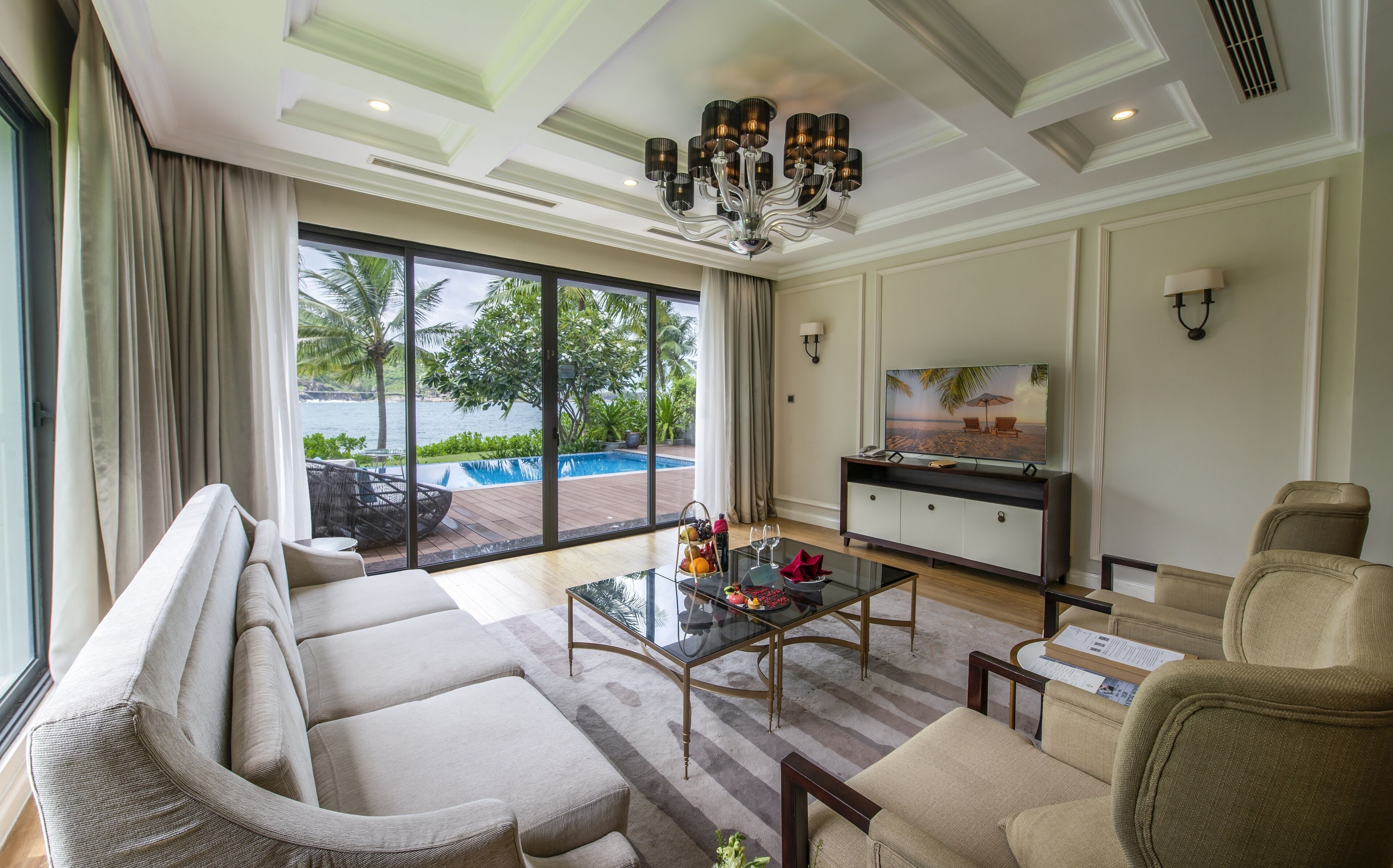 Ein geräumiges Wohnzimmer mit einem großen Fenster, das einen Blick auf einen Pool und Palmen bietet. Es gibt ein Sofa, einen Couchtisch mit Obst und Weingläsern sowie einen Fernseher auf einem Schrank.