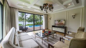 Ein geräumiges Wohnzimmer mit einem großen Fenster, das einen Blick auf einen Pool und Palmen bietet. Es gibt ein Sofa, einen Couchtisch mit Obst und Weingläsern sowie einen Fernseher auf einem Schrank.