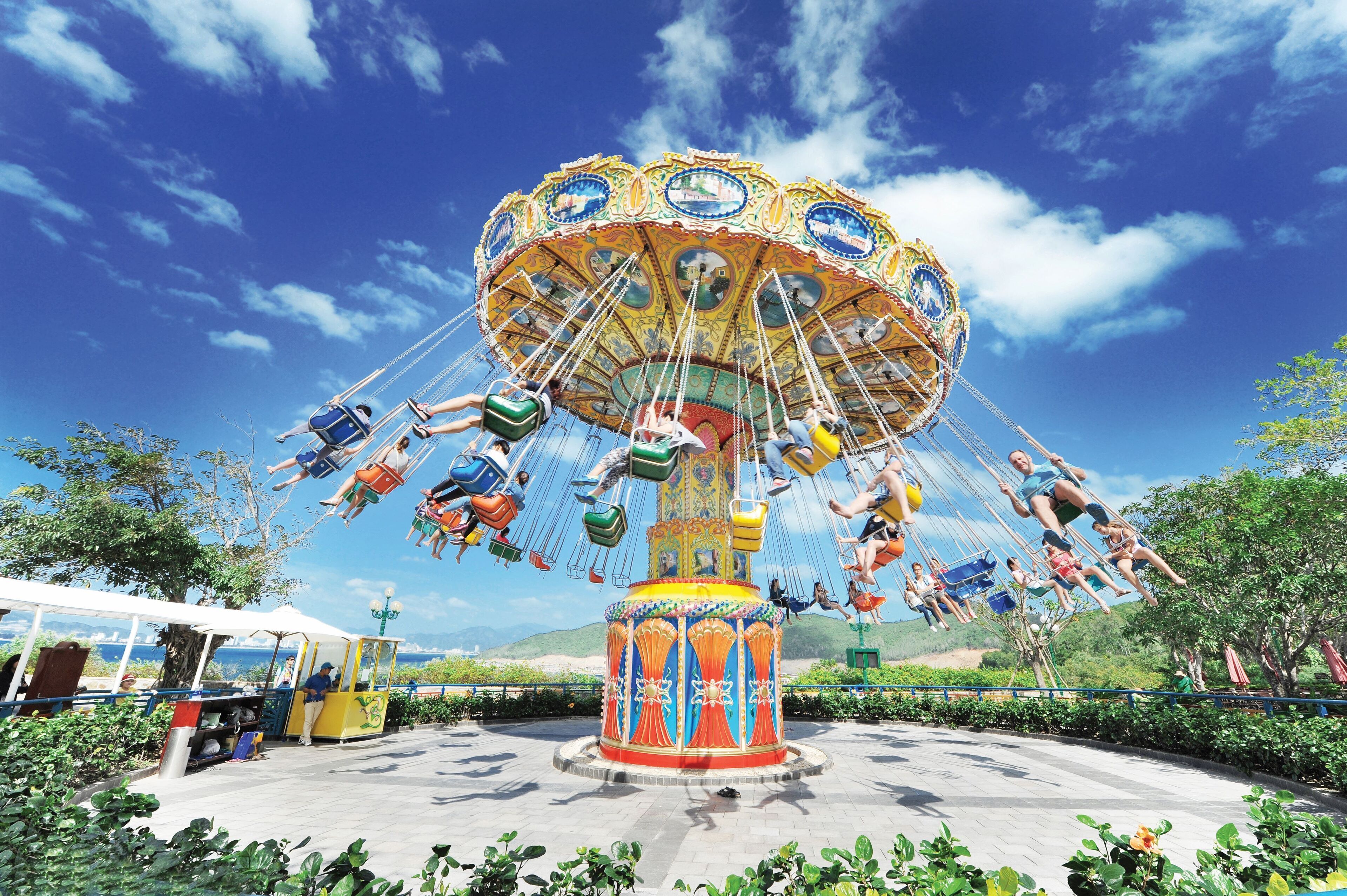 A carousel with multiple seats and a central column, surrounded by greenery and a clear sky.