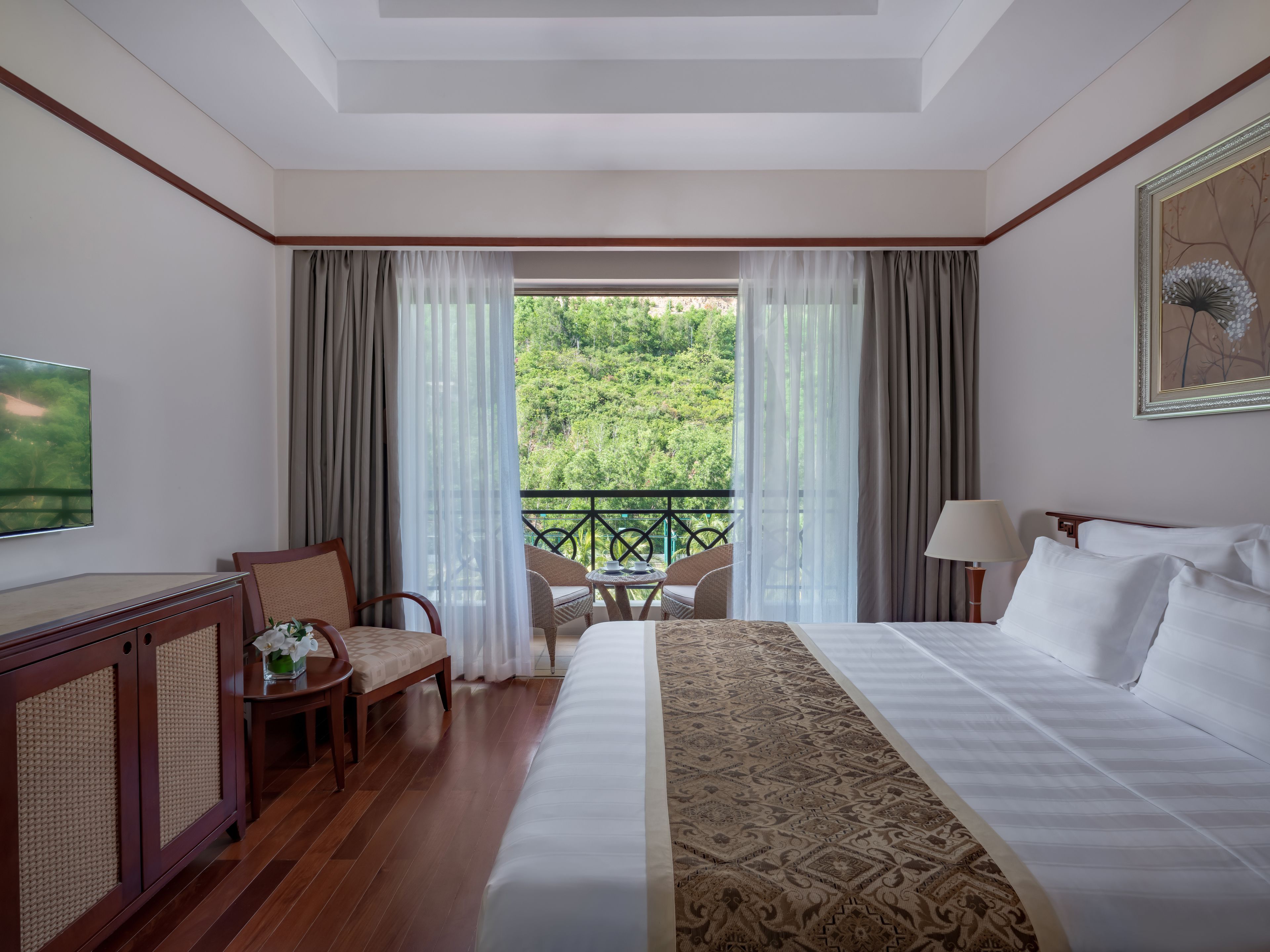 Habitación de hotel con una cama grande, televisión, escritorio con silla, mesita con silla y balcón con vistas a zonas verdes.