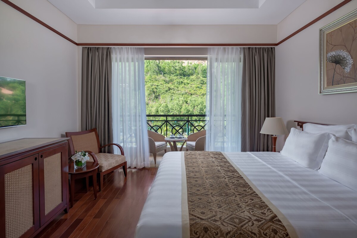 Habitación de hotel con una cama grande, televisión, escritorio con silla, mesita con silla y balcón con vistas a zonas verdes.
