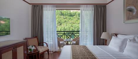 Habitación de hotel con una cama grande, televisión, escritorio con silla, mesita con silla y balcón con vistas a zonas verdes.