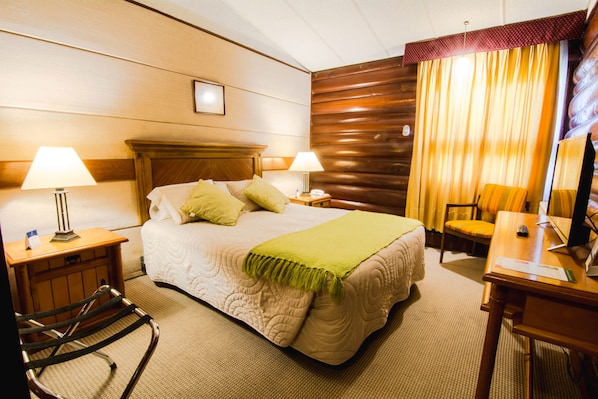 Habitación de hotel con una cama grande, dos mesitas de noche con lámparas, un cabecero de madera, un escritorio con silla y un ventanal con cortinas.