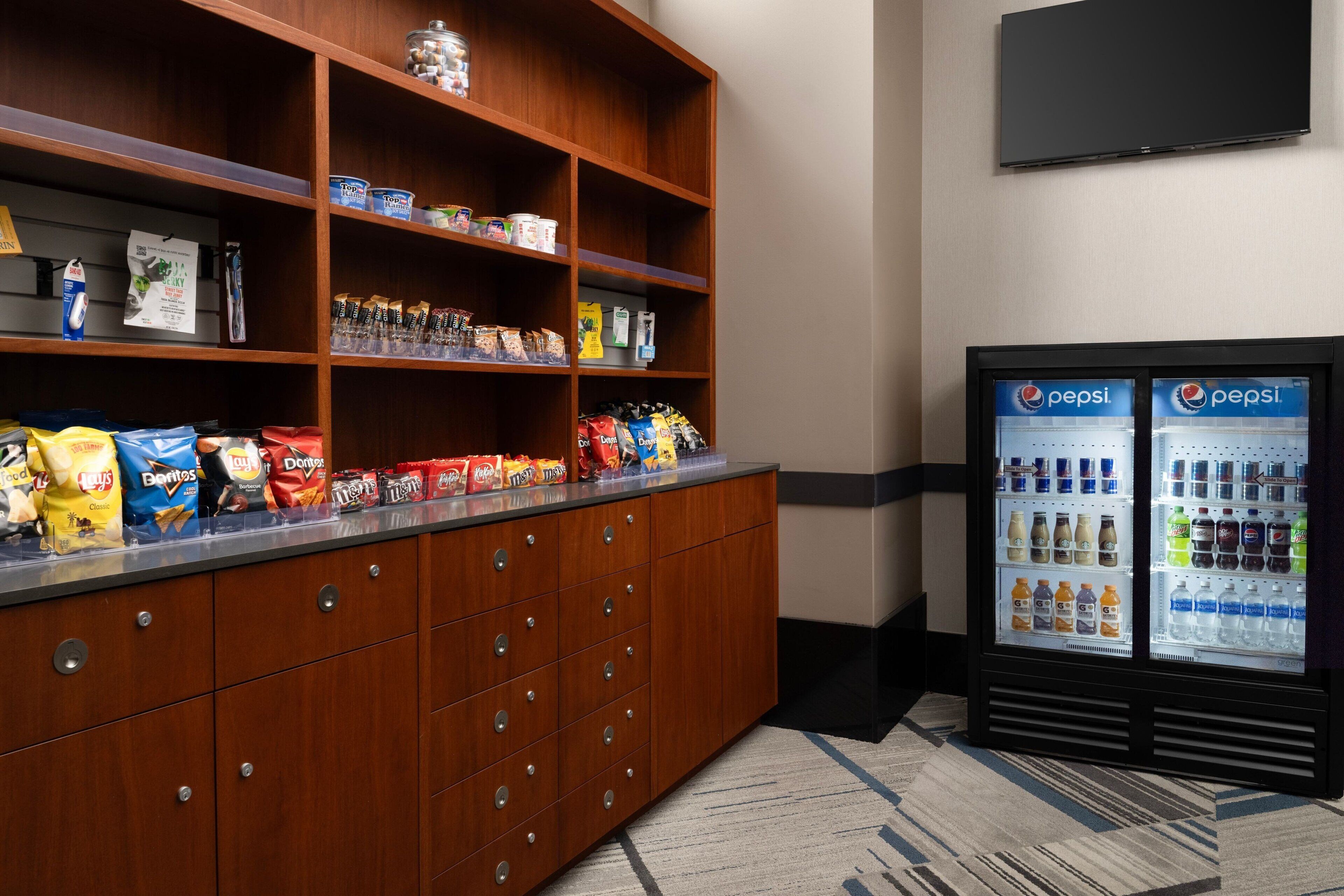 Un bar de aperitivos con varias opciones de papas fritas y bocadillos, un refrigerador con productos Pepsi, y un televisor montado en la pared.
