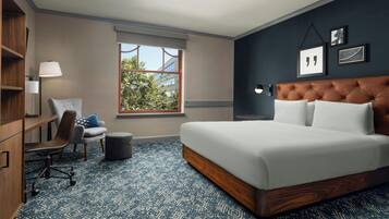 A hotel room with a large bed, a desk, a chair, a window, and a wall with framed pictures.