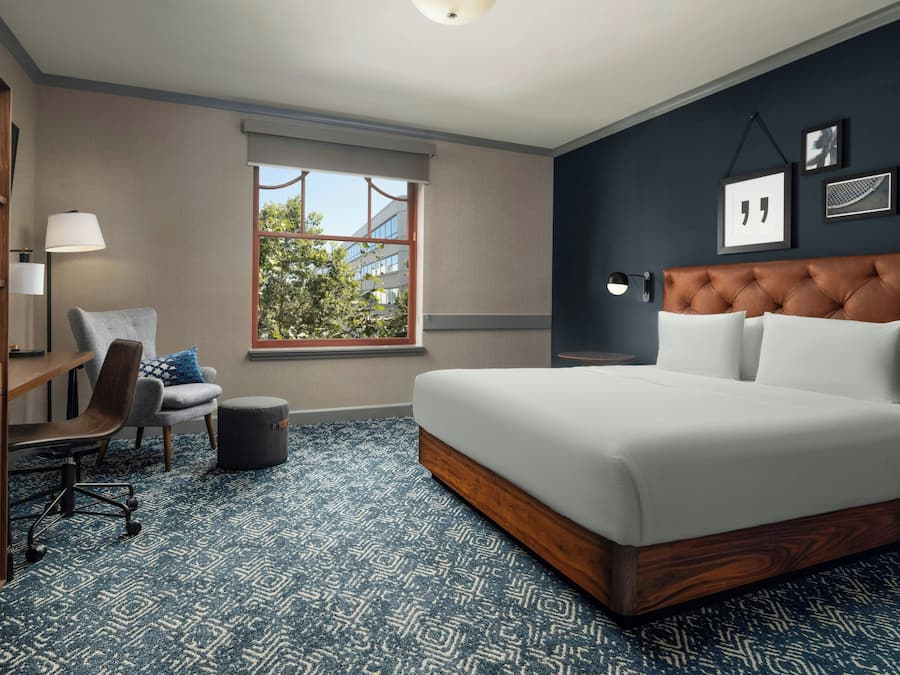 A hotel room with a large bed, a desk, a chair, a window, and a wall with framed pictures.