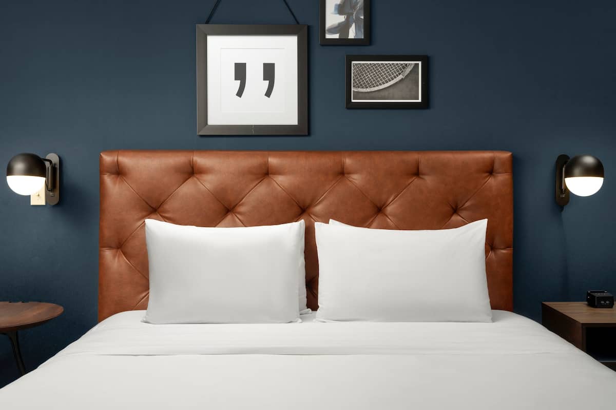 Une chambre à coucher avec une tête de lit capitonnée, deux oreillers blancs, un lit avec du linge de lit blanc, une table de chevet ronde et deux lampes fixées au mur.