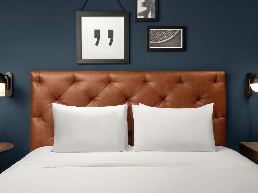 Un dormitorio con cabecera capitonada, dos almohadas blancas, una cama con ropa de cama blanca, una mesita redonda y dos lámparas fijadas a la pared.