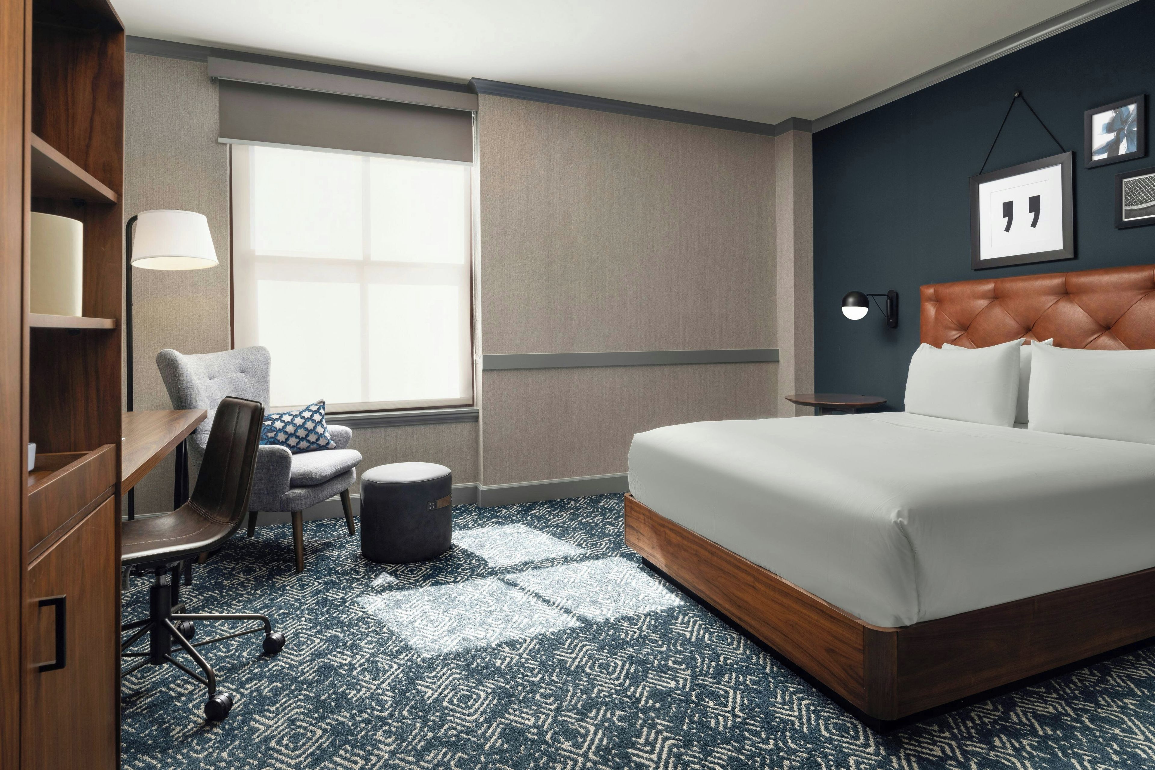 A hotel room with a large bed, a desk with a chair, a small ottoman, a window with blinds, a wall-mounted lamp, and a framed quote on the wall.
