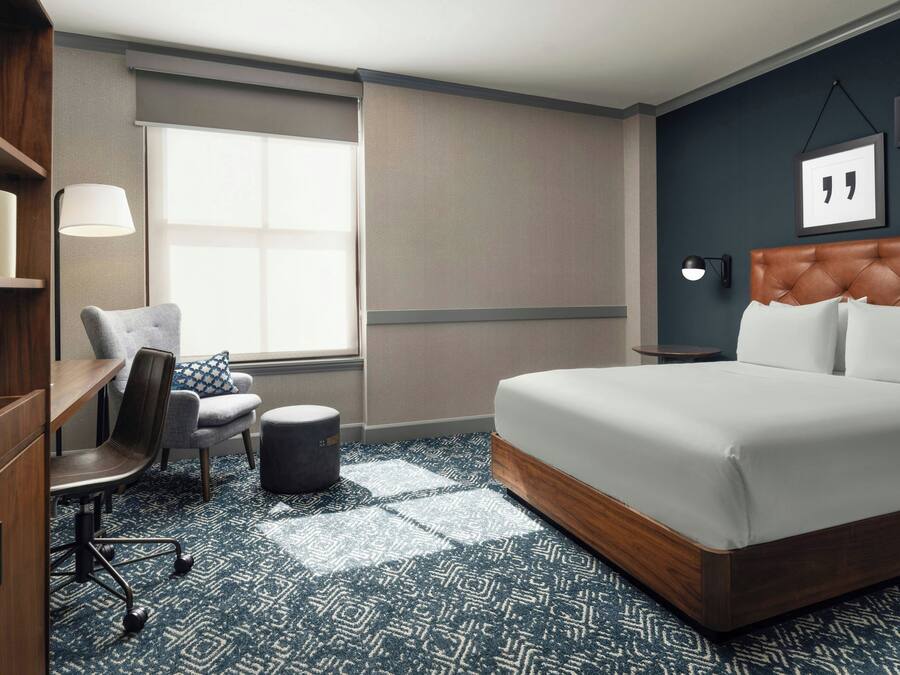A hotel room with a large bed, a desk with a chair, a small ottoman, a window with blinds, a wall-mounted lamp, and a framed quote on the wall.