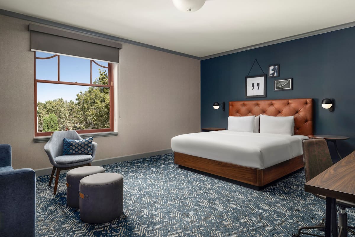 Une chambre à coucher avec un grand lit, une chaise, des poufs, un bureau et une fenêtre donnant sur des arbres.