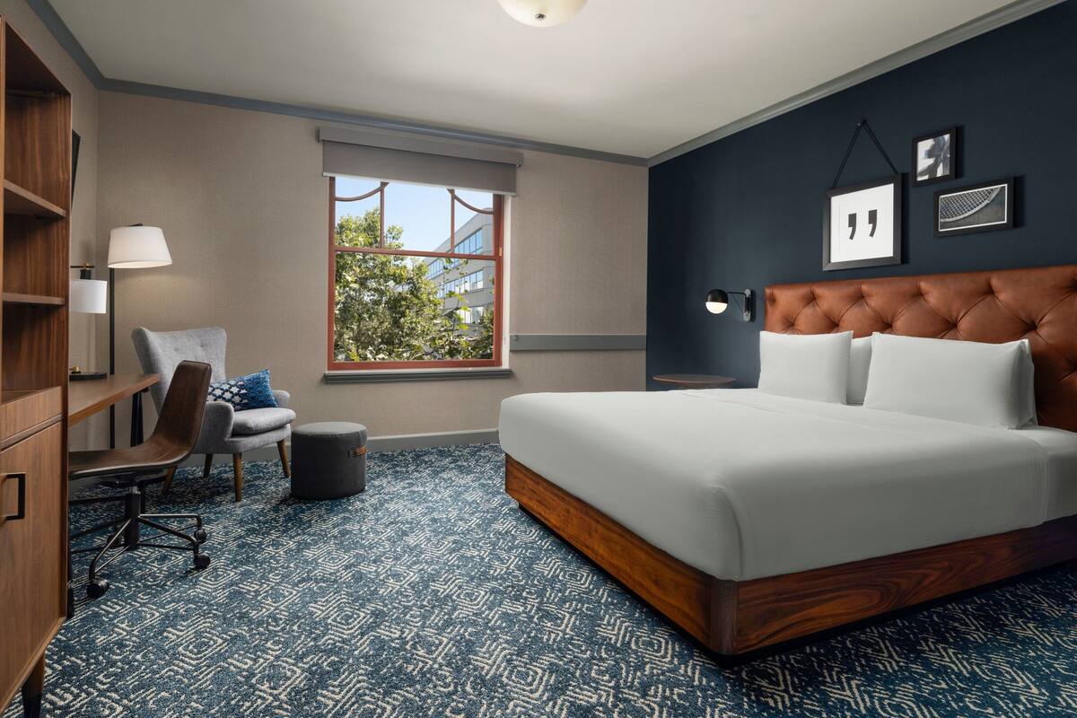 Une chambre d’hôtel avec un grand lit, un bureau avec une chaise, un petit tabouret et une fenêtre donnant sur des immeubles et des arbres.