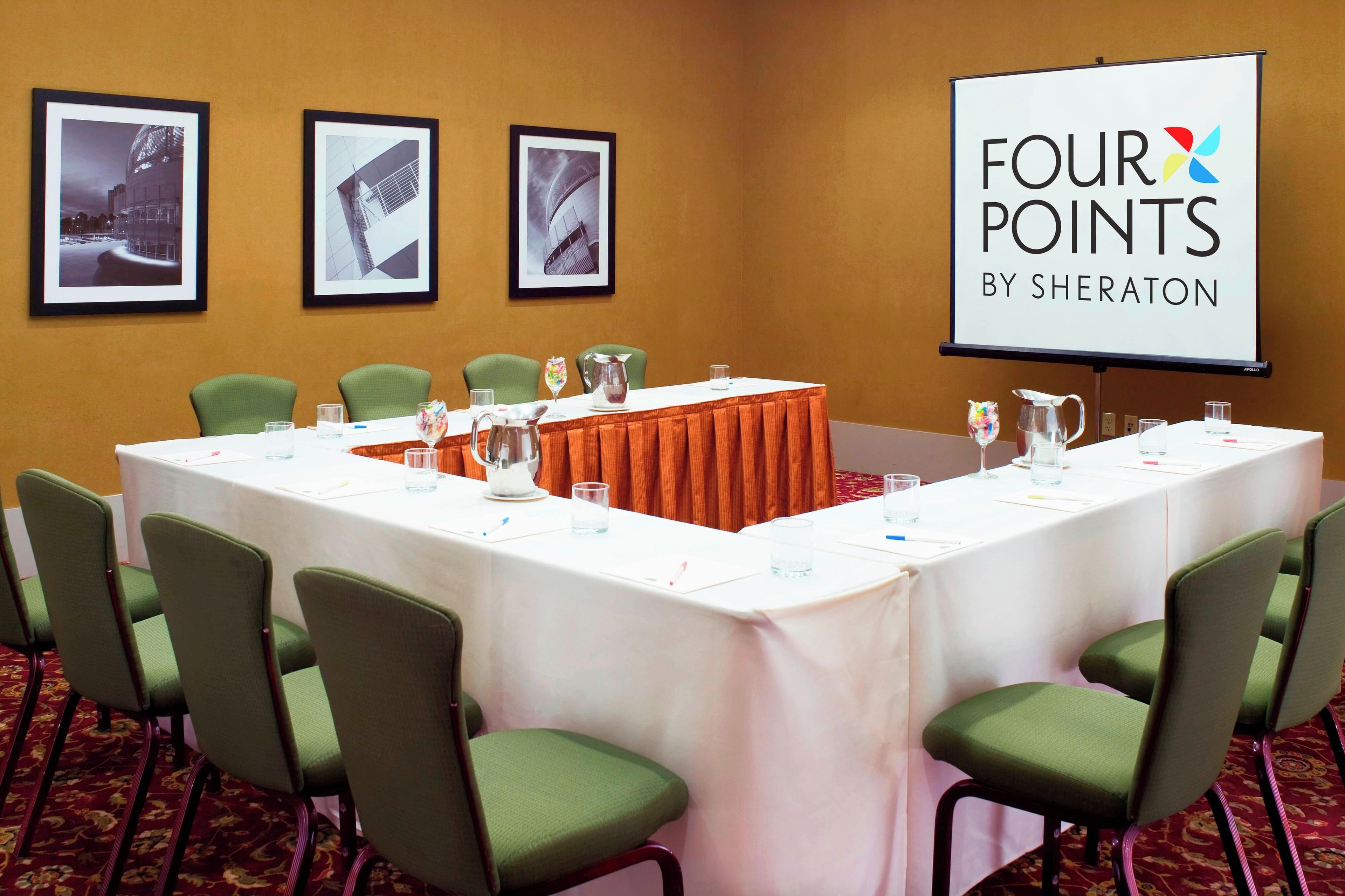 A conference room with a long table, green chairs, a projector screen, and framed pictures on the wall.