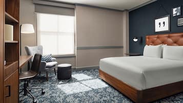 Habitació d'hotel amb un llit gran, un escriptori amb una cadira, un otomà petit i rodó, una làmpada i una finestra amb persianes.