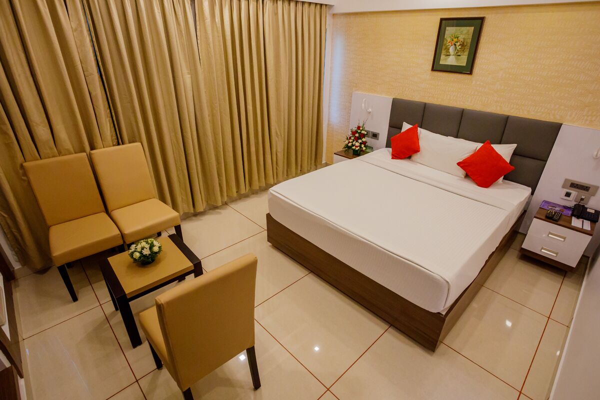 Habitación de hotel con una cama, dos sillas, una mesita con un florero, una mesita de noche y una cortina.