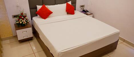 Una cama bien hecha con almohadas blancas y rojas, una mesita de noche con un teléfono y un pequeño jarrón de flores, y un mueble con cajones.