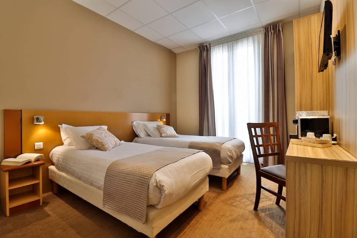 A hotel room with two beds, a desk, a chair, a TV, and a window with curtains.