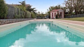 Una piscina rectangular amb superfície d'aigua blava cristal·lina, envoltada per una paret de pedra i una tanca de fusta. Al fons, hi ha un edifici petit amb teulada vermella, una porta i finestres.