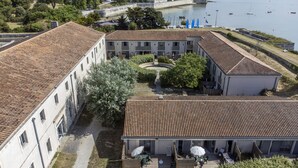 Un pati amb un edifici amb teulada de rajola, envoltat de vegetació i una massa d'aigua amb embarcacions.