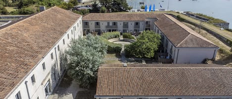 A courtyard with a building featuring a tiled roof, surrounded by greenery and a body of water with boats.