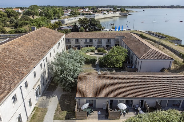 A courtyard with a building featuring a tiled roof, surrounded by greenery and a body of water with boats. - Pierre & Vacances Ill d'Aix Résidence Fort de la Rade (Ile-d'Aix)