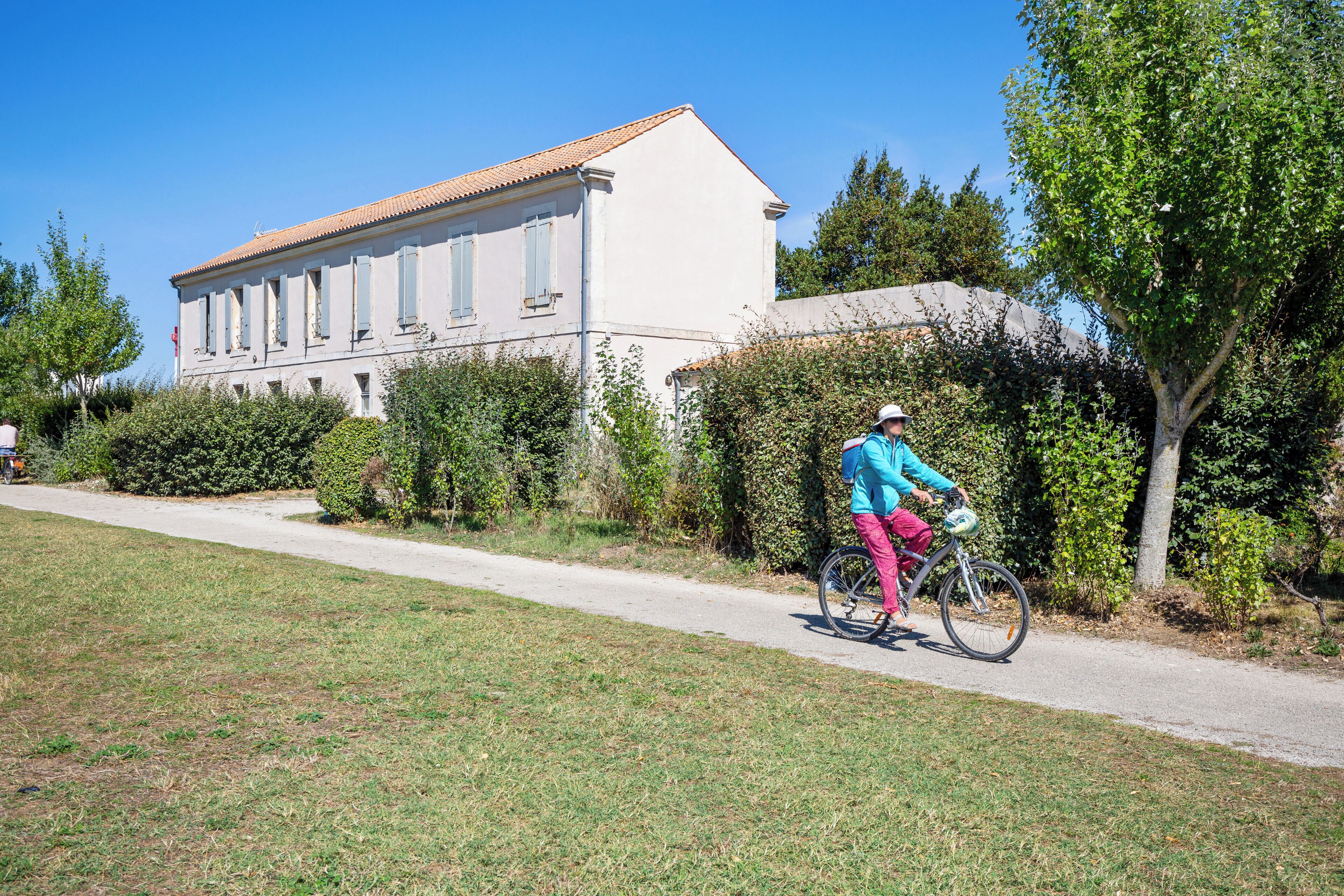 Uma pessoa andando de bicicleta em um caminho pavimentado, ao lado de um edifício com telhado telhado e várias janelas.