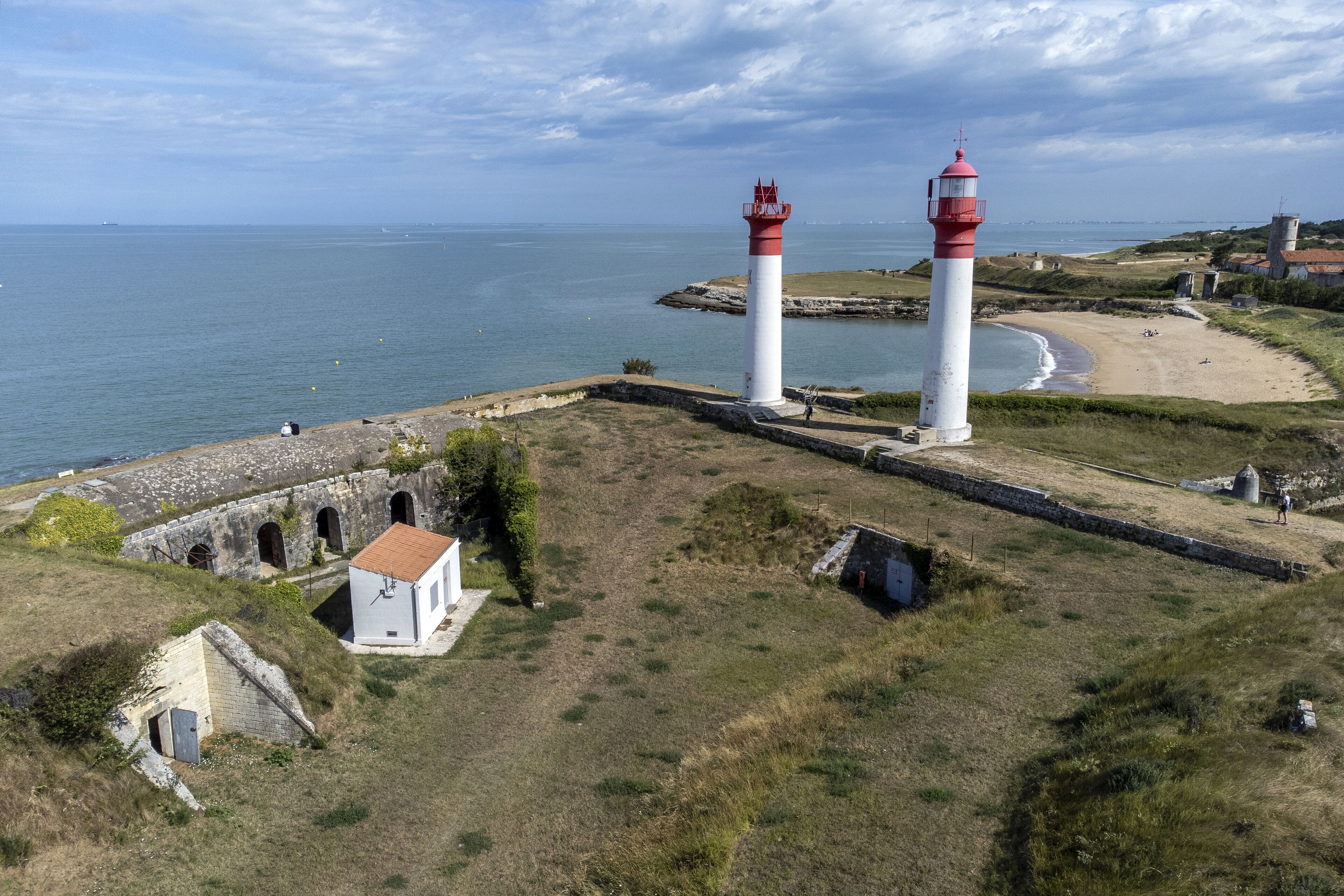 A coastal lighthouse with a white and red structure, a stone building with a red roof, and a sandy beach in the background.