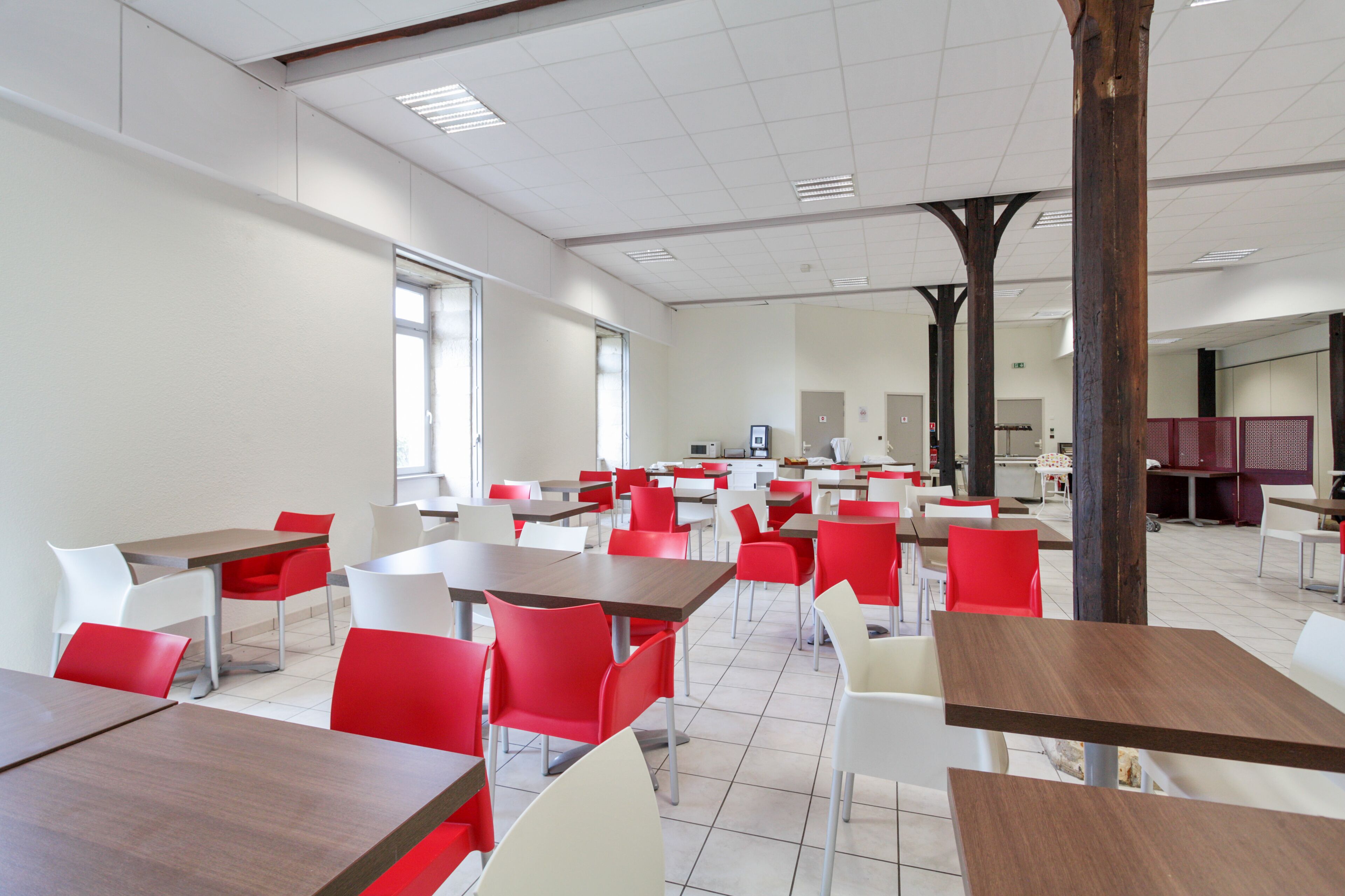 A cafeteria with multiple tables and chairs, featuring a mix of red and white seating, and a tiled floor.