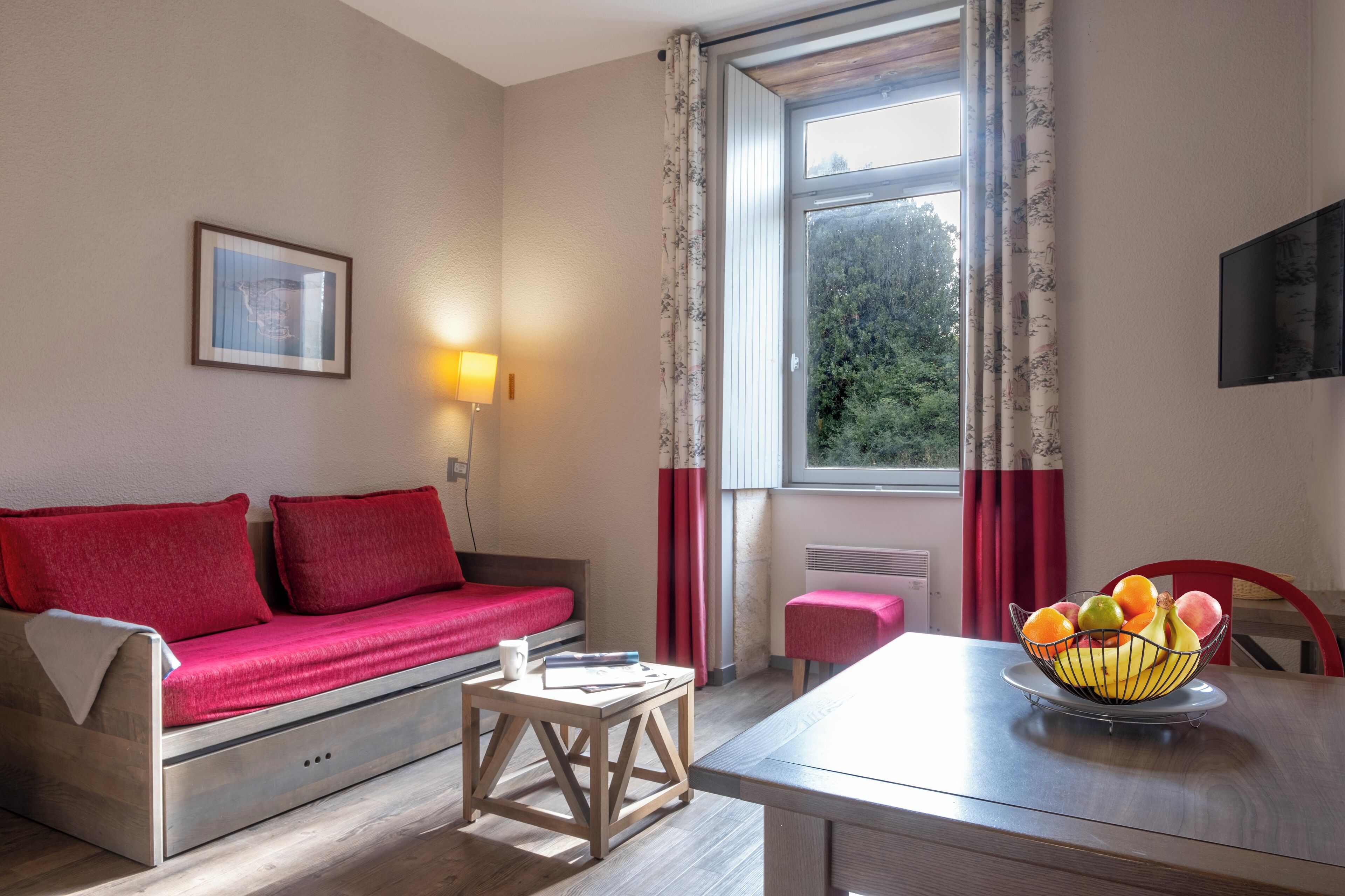Een kamer met een bank, een tafel met een schaal fruit, een lamp en een raam met gordijnen.