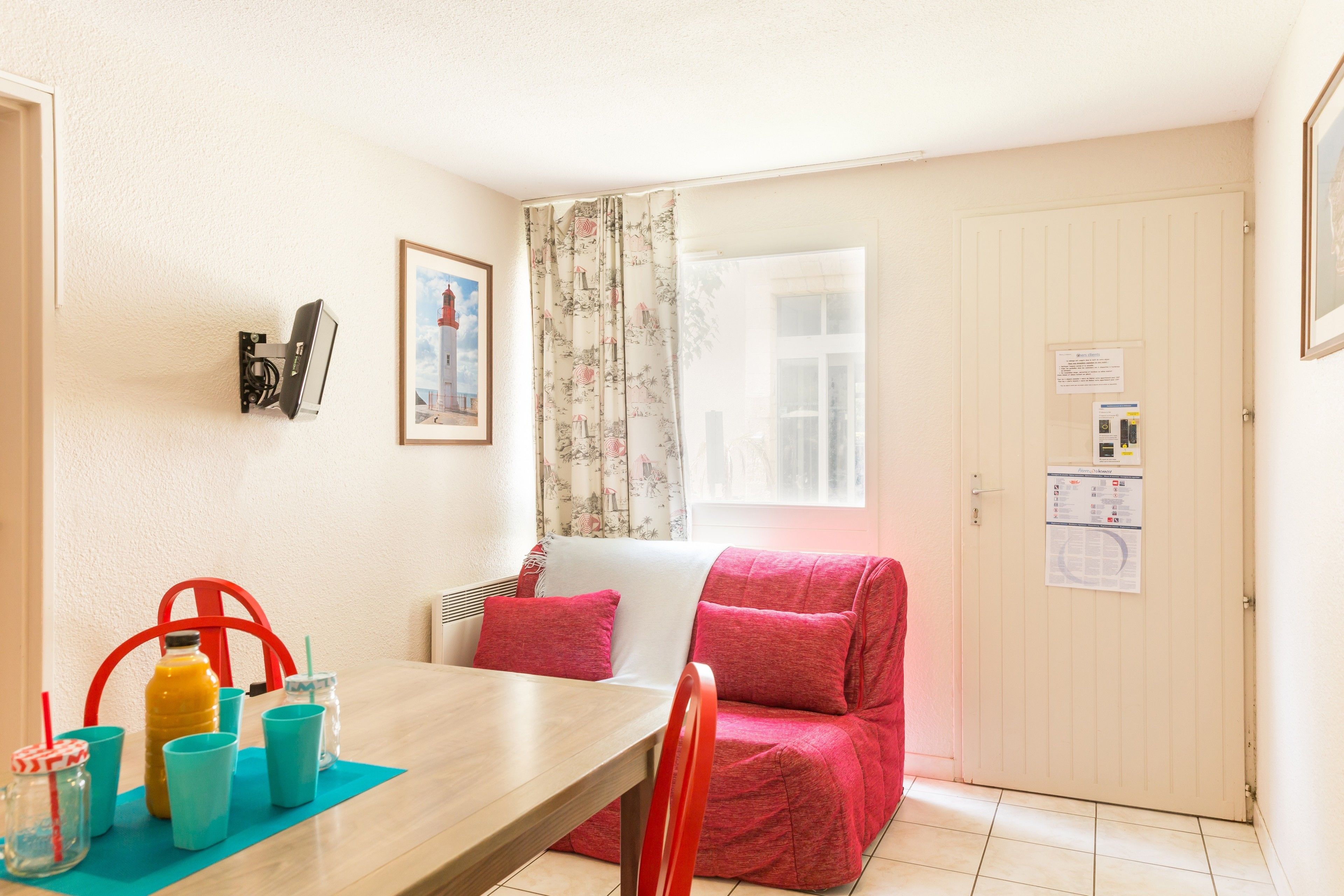 A compact room with a dining table, red and white sofa, and a window with floral curtains.