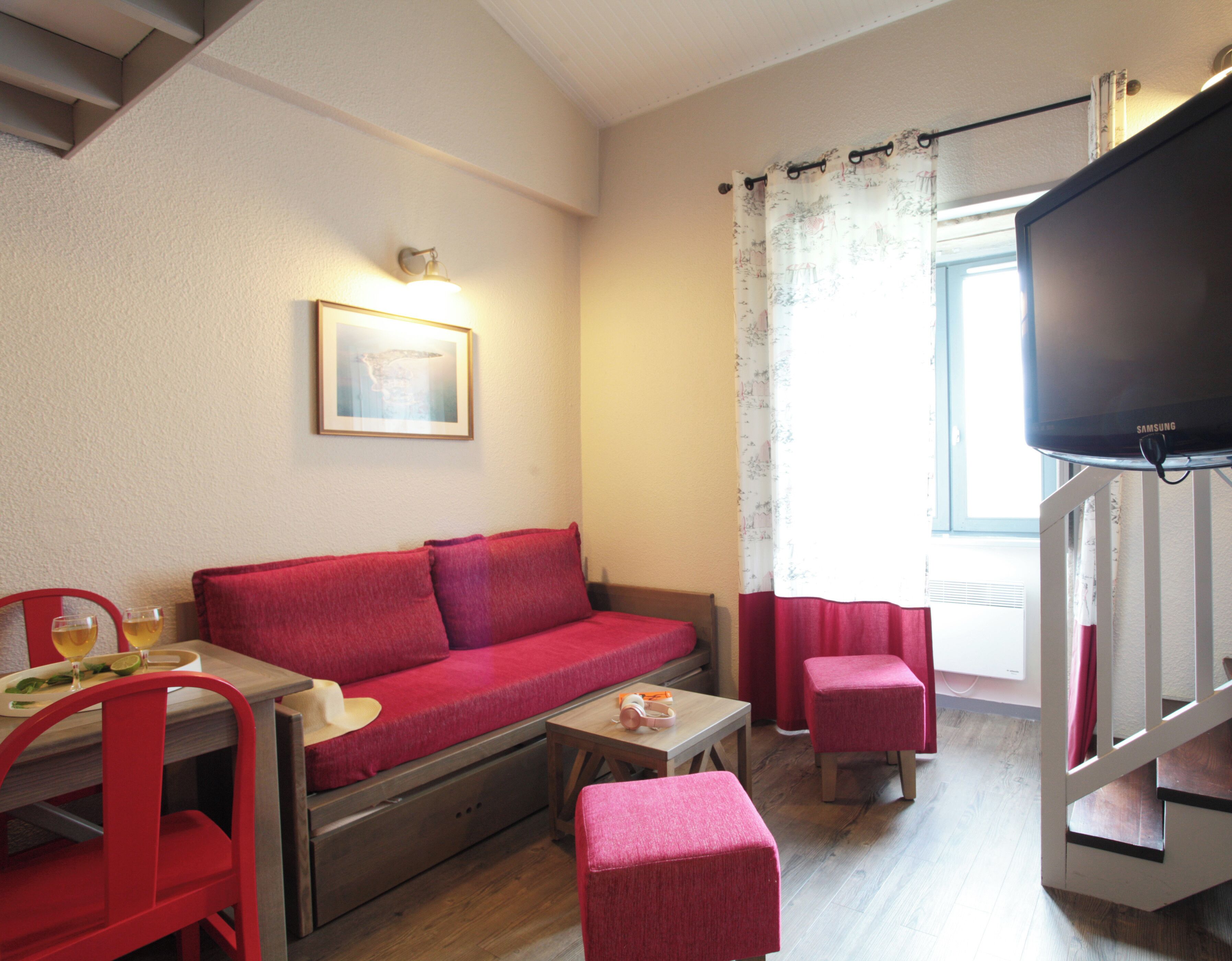 A compact living space with a red sofa, a small dining table, a TV, and a window with curtains.
