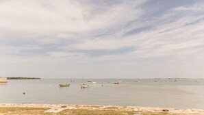 Escena costanera amb vaixells sobre aigua tranquil·la, una platja sorrenca i una línia de costa llunyana.