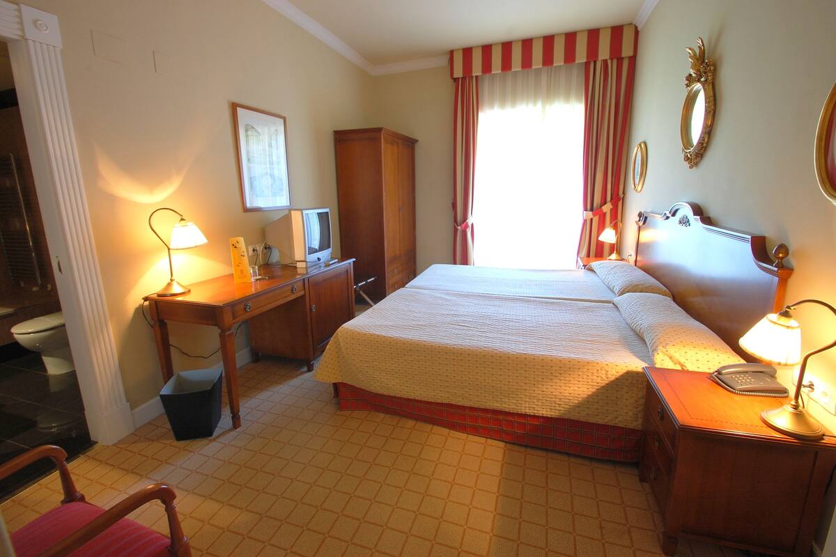 Habitación de hotel con cama, mesitas de noche, escritorio, silla, televisor y baño con inodoro y lavamanos.