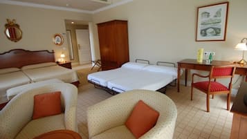 Una habitación de hotel con dos camas individuales, una cama doble, un armario de madera, un escritorio con una silla y un cuadro en la pared.