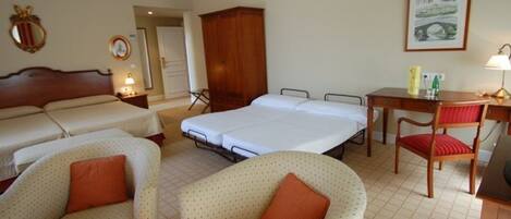 Quarto de hotel com duas camas de solteiro, uma cama de casal, um guarda-roupa de madeira, uma mesa de trabalho com cadeira e um quadro na parede.