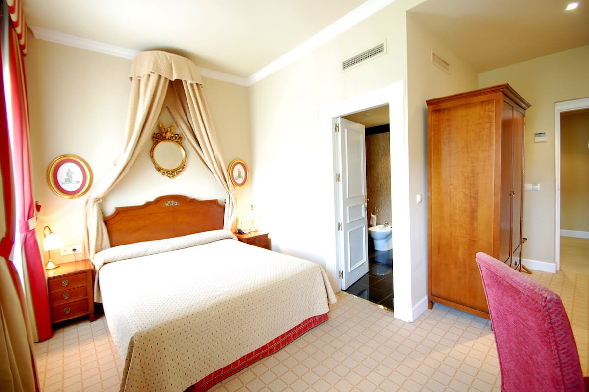 Habitación de hotel con una cama grande, un armario de madera, un baño con inodoro y lavabo, y una silla roja.
