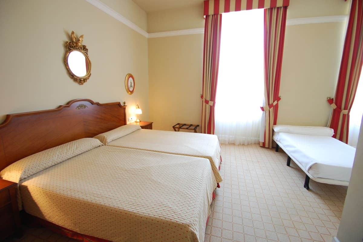 Habitación de hotel con dos camas, cabecero de madera, mesita de noche con lámpara, una pequeña mesa y ventana con cortinas a rayas.