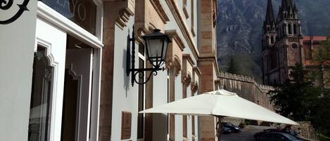 A building with a white facade, black wrought iron window frames, and a large umbrella providing shade. There is a street with cars and a mountain in the background.