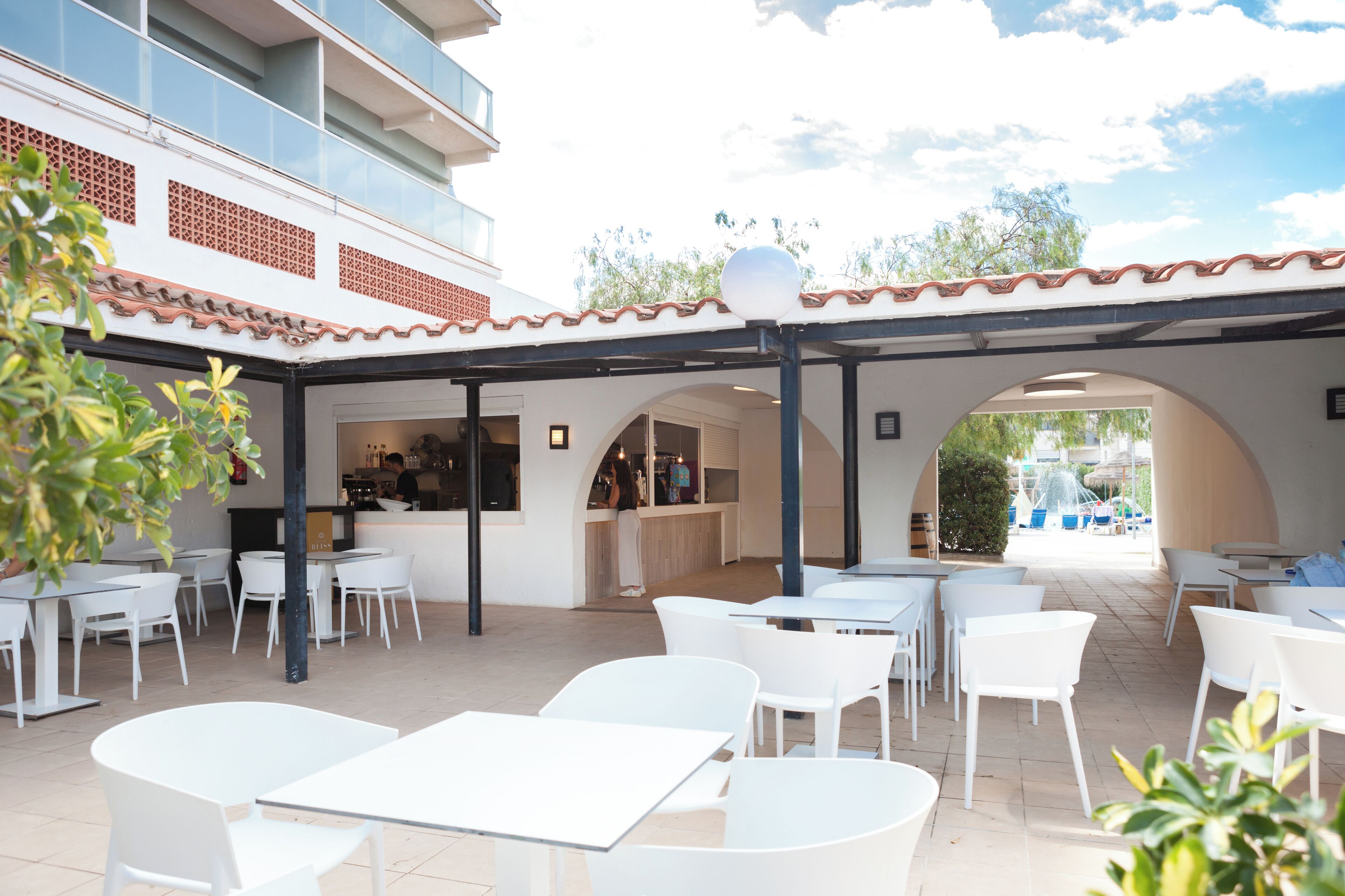 An outdoor patio area with white tables and chairs, a bar area, and a view of a pool and fountain.