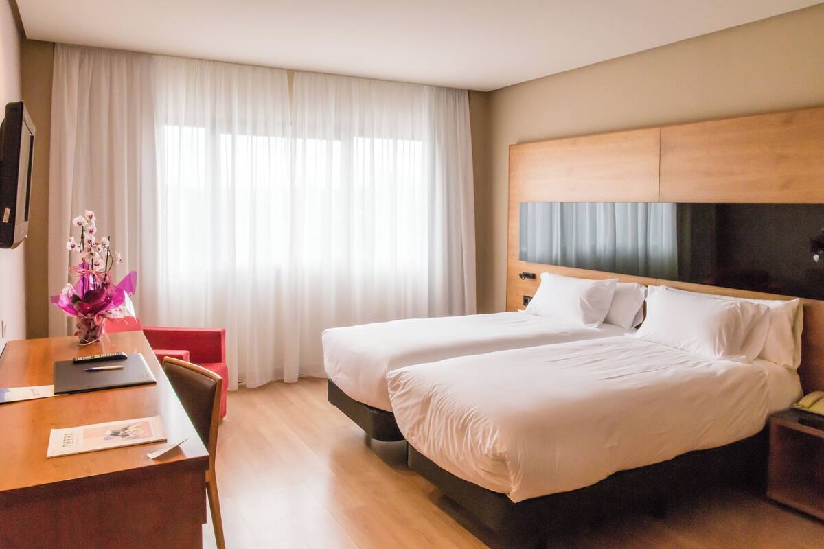 A hotel room with two beds, a wooden desk, a chair, a TV, and a window with white curtains.