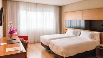 Ett hotellrum med två sängar, ett skrivbord i trä, en stol, en tv och ett fönster med vita gardiner.