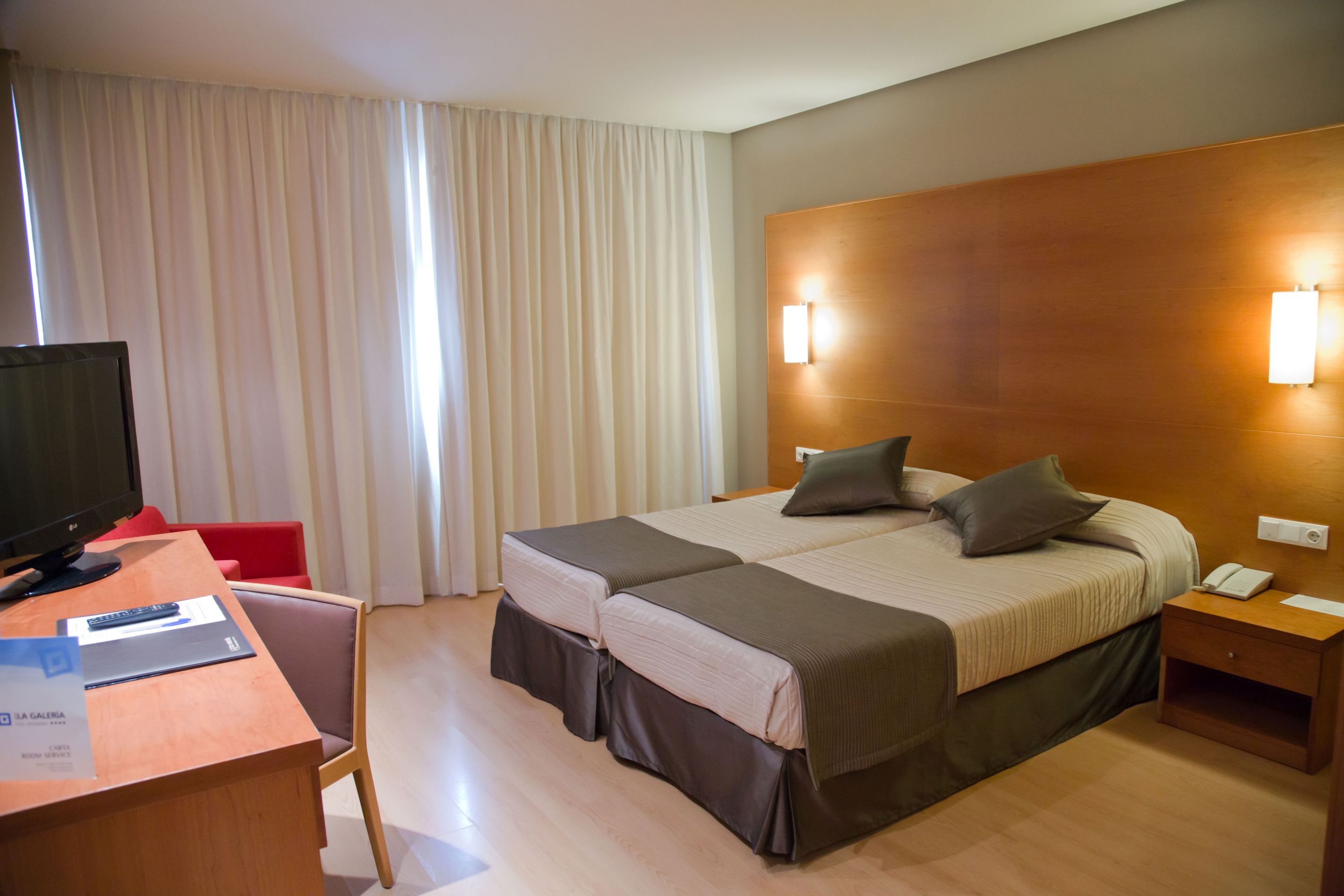 Une chambre d’hôtel avec une tête de lit en bois, deux lits avec la même literie, un bureau avec une télévision et un fauteuil rouge.